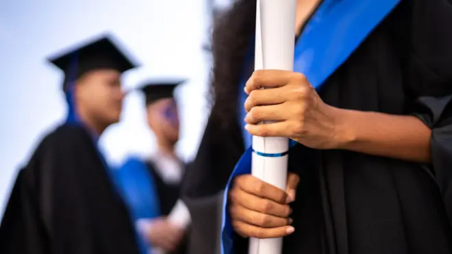 Elinde rulo halinde diploma tutan üniversite mezuniyet kıyafeti giyen bir kadın ve arka planda mezuniyet kepli iki erkek öğrenci