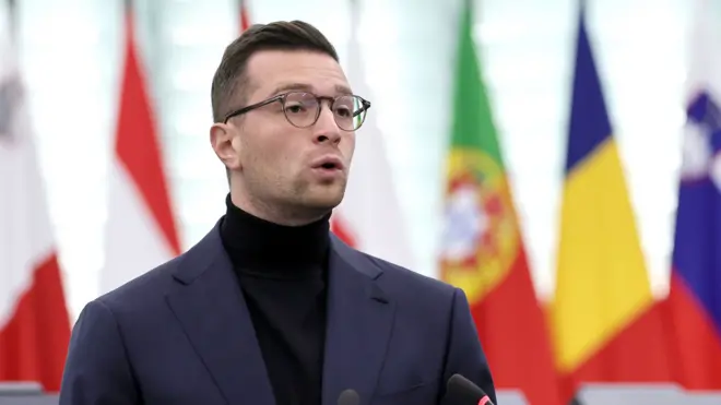 Bardella speaks while wearing a dark turtleneck and suit in front of European flags, at the European Parliament in October.