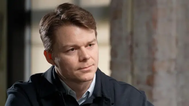 Palantir UK boss Louis Mosley speaking to the BBC. He is wearing a dark jacket with a button up jumper and is sitting in front of a grey wall. 
