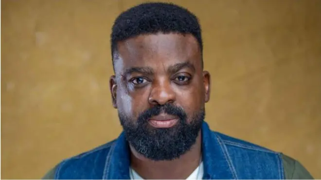 Aworan oṣere tiata, Kunle Afolayan. O wọ aṣọ alawọ aro pẹlu awọtẹnu funfun. Irun ori ati ti ààgbọ̀n rẹ pọ ninu aworan naa, bẹẹ lo n wo ọọkan.
