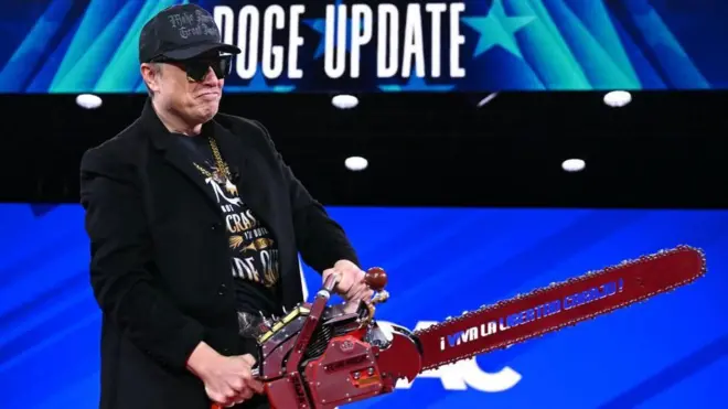Elon Musk, wearing a hat and glasses, holds a chainsaw