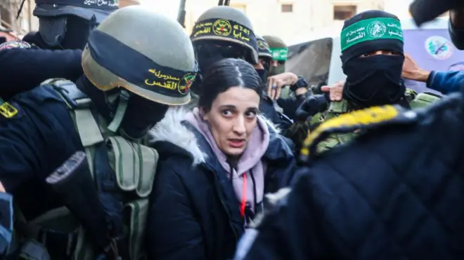 أربيل يهودا بلباس شتوي وجاكيت تقف بين مقاتلي الجهاد الإسلامي وحماس لحظة تسليمها وسط حشود في خان يونس في قطاع غزة