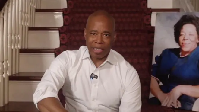 Эрик Адамс в белой рубашке без пиджака сидит на ступеньках лестницы, покрытой ковром, рядом с прислоненным к перилам портретом женщины