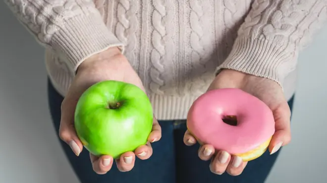 Pessoa oferece uma maçã verde em uma das mãos e um donut rosa na outra.