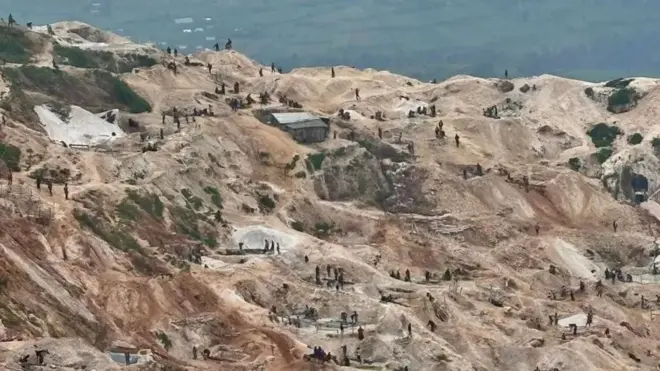 Vue aérienne des activités minières à la mine de Rubaya, où le terrain est vallonné et légèrement en terrasses. La terre nue est de couleur brun clair, avec par endroits des nuances plus foncées tirant vers l'orange. On aperçoit au loin plusieurs groupes de mineurs en train de nettoyer le minerai. On distingue une cabane en tôle ondulée. Au loin, on aperçoit des champs verdoyants.