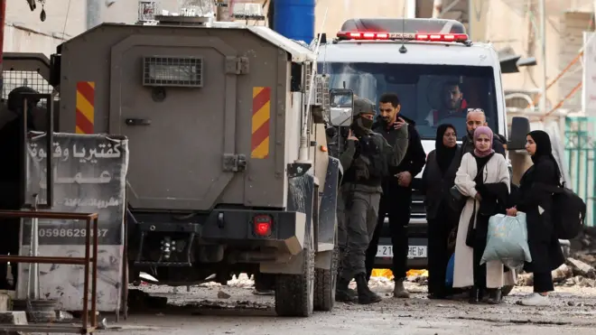 صورة تظهر عائلات نازحة من مخيم جنين، إلى جانب مركبة عسكرية إسرائيلية.