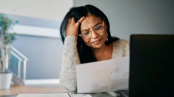Uma mulher estressada está sentada à mesa em casa, lendo um documento ao lado do laptop. Ela parece preocupada e tensa, com a cabeça apoiada nas mãos enquanto administra suas contas pessoais.