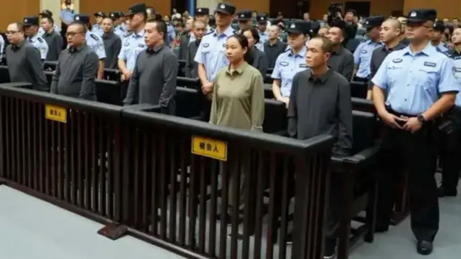 Las personas están de pie en fila en un tribunal, rodeadas por agentes de seguridad.