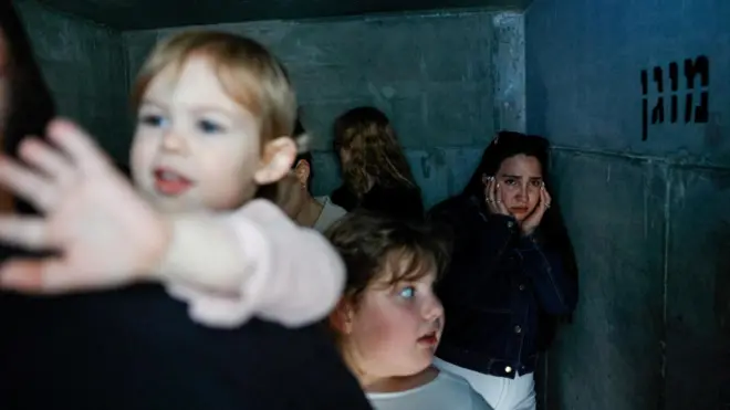 Невелике укриття з бетонними стінами, де перебуває група молодих жінок і дітей. Тісний простір освітлений слабким холодним світлом, одна з дівчат дивиться стривожено в камеру, притискаючи руки до обличчя.