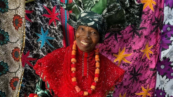 Maman Nike fait face à l'objectif, vêtue d'une robe rouge vif ornée de perles et d'un grand foulard vert à motifs, rehaussée d'un collier de grosses perles rouges et orange. Derrière elle se déploie une sélection de textiles aux motifs floraux et étoilés, dans des tons de noir, rose, violet, blanc et autres.