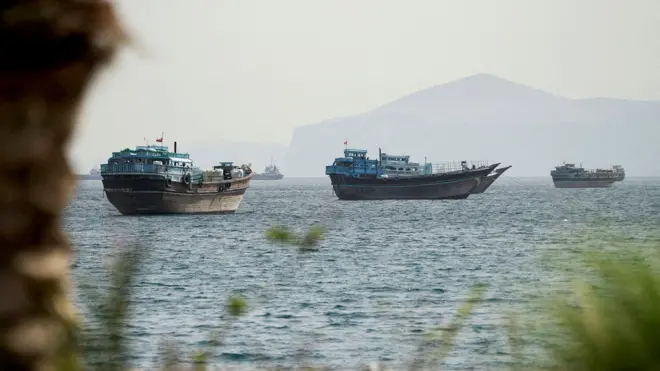 Boats atop the water pictured in a view of the Strait of Hormuz on a hazy day in mid-May with rock and plantation bokeh in the foreground and mountains in the background, taken from Oman on 22 May.