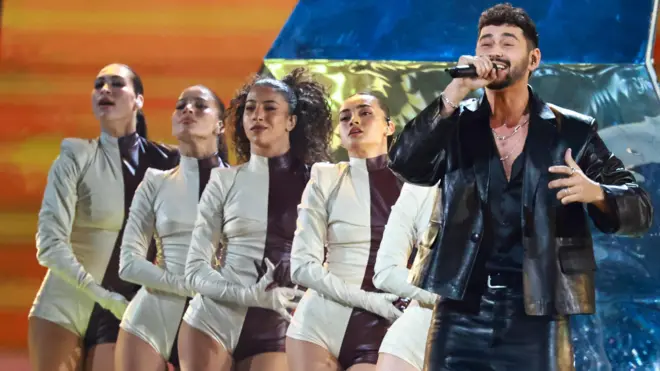 Noam Bettan Eurovision yarışmasında finale kaldı. Baştan aşağı siyah deri kıyafet giymiş bir şekilde şarkı söylüyor. Arkasında beş kadın sanatçı var.