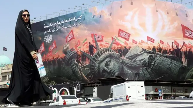 自由の女神が倒れ、その後ろで人々が赤い旗やイラン国旗を掲げている大きな絵が、建物に掲示されている。そのそばを黒いベールで身を包んだ人が歩いている