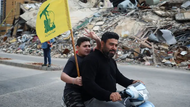 Two people ride on a moped, while one holds a Hezbollah flag and holds two fingers in the air, on a street in Beirut in front of a huge pile of rubble where a building once stood, taken on Friday.
