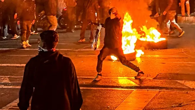 A masked protester raises his hand in font of a burning fire in the street at night