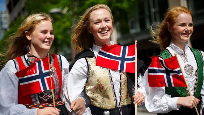 Üç tane kadın norveç bayrakları ile yürüyor. Hepsi sarışın ve gülüyorlar.