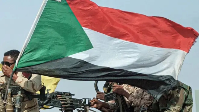 العلم السوداني يرفرف فوق مركبة مثبت عليها مدفع رشاش يحمله جنود من قوات الدعم السريع.
