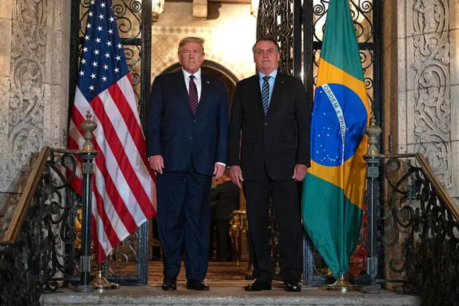 Trump e Bolsonaro em pé, com olhar sério, em frente a porta sofisticada