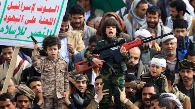 Толпа на митинге. В центре на плечах взрослых двое детей в камуфляже с макетами пистолета и автомата