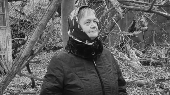 Чорно-білий портрет літньої жінки в куртці та хустці з квітами на тлі поламаних дерев закинутого подвірʼя