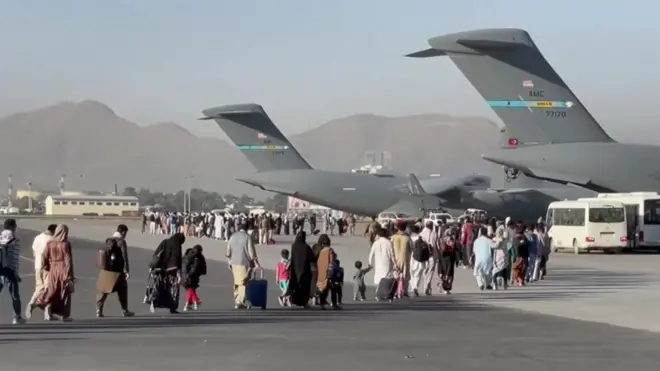 People carrying luggage walk across an airport tarmac towards large US military transport aircraft during an evacuation