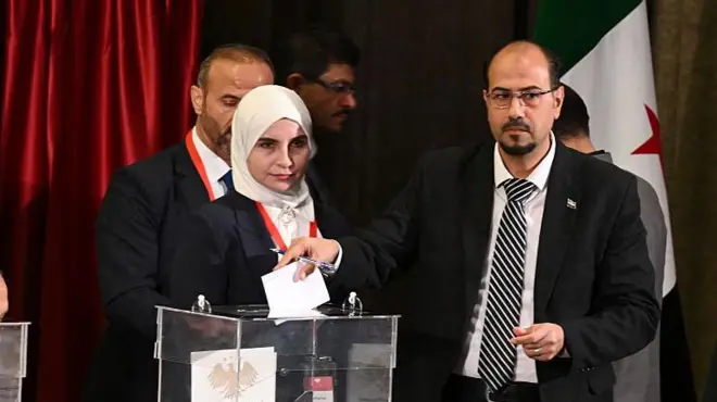 Bir kadın ve erkek yan yana camdan sandığın başındalar. Başörtülü kadın kameraya bakıyor, erkek oy atıyor.