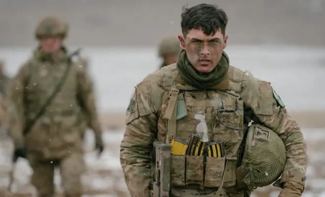 Karlı bir arka planda bir İngiliz komandosu kameraya bakarak yürüyor. Arkasında flu durumda başka askerler de var.