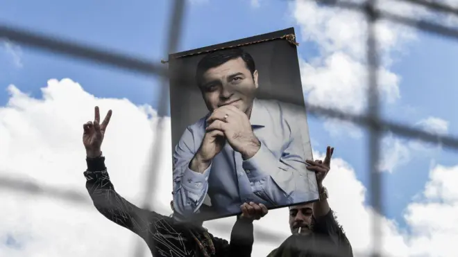 İki adam Selahattin Demirtaş'ın posterini kaldırıyor. elleriyle barış işareti yapıyorlar. Arkada mavi gökyüzü.