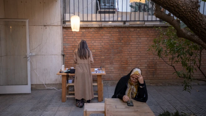 تصویر زنی میانسال با چادر مشکی در یک کافه مشغول نگاه کردن به گوشی موبایلش. در پشت او دختری بدون حجاب ایستاده است.