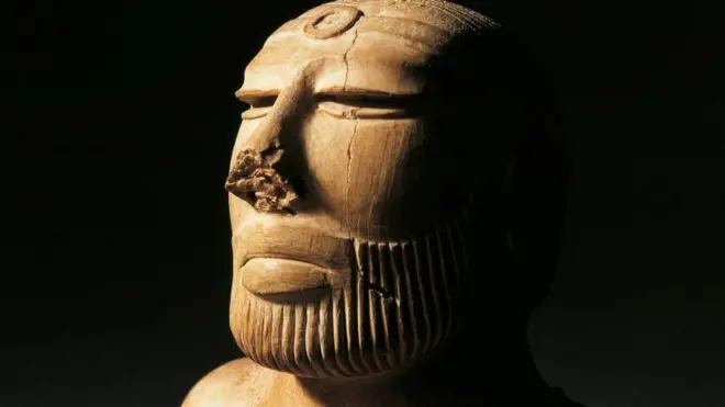 Uma estátua em close do rosto, com barba bem aparada, iluminada por luz amarelada contra um fundo preto. A escultura tem um círculo entalhado na testa, e o nariz parece parcialmente desgastado