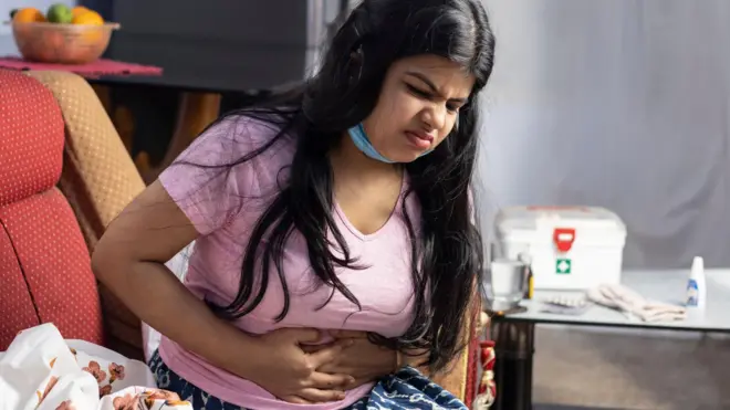 A mid adult Indian woman wearing surgical nose mask, sitting on sofa, feeling stomachache, health care concept