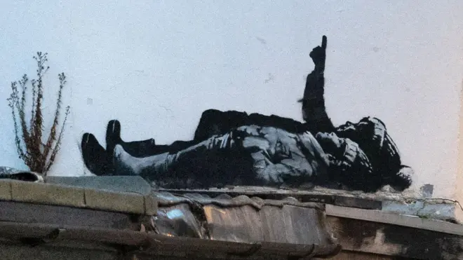 The black and white street art depicts two children lying on their backs and pointing upwards. The child nearest the camera is wearing a wooly hat with hands tucked into their jacket pockets. Behind them a larger person is pointing upwards in a bobbly hat (possibly a Santa type hat) also wearing boots.