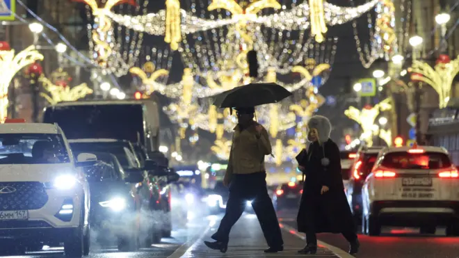 Двое людей переходят улицу, украшенную новогодними гирляндами