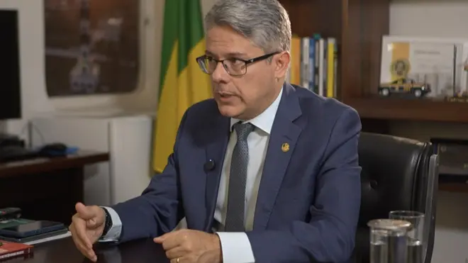 Alessandro Vieira está sentado em uma cadeira em seu gabinete. Ele usa um terno azul com uma gravata cinza a blusa branca, além de óculos de grau.