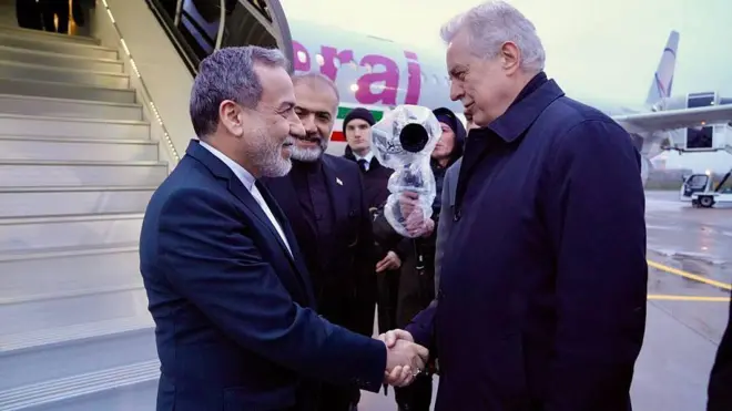 Двое мужчин пожимают друг другу руки у трапа самолета, на борту которого изображен флаг Ирана.