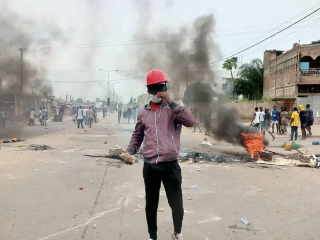 Un jeune manifestant avec casque rouge, visage caché et une pierre à la main droite. derrière lui des pneus brûlés avec des flammes, de la fumée noire et d'autres manifestants.