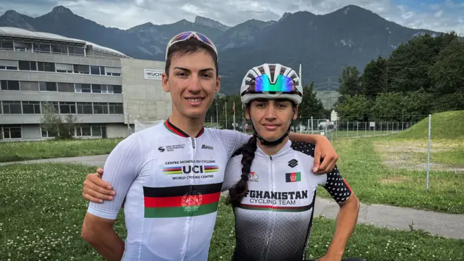 Yulduz and Fariba Hashimi standing arm in arm in cycling gear in front of their training centre and some mountains