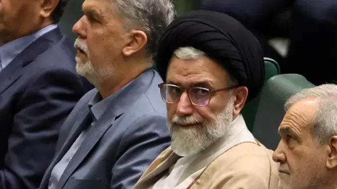 緑色の椅子に、複数の白髪の男性が座り、眼鏡をかけ、クリーム色の法衣と黒いターバンを身に着けた白いひげの男性がカメラの方を見ている