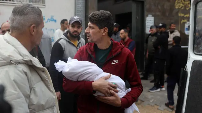 Orta yaşlı bir adam kucağında beyaz kefene sarılı bir bebek tutuyor, çevresinde üzgün duran erkekler var.