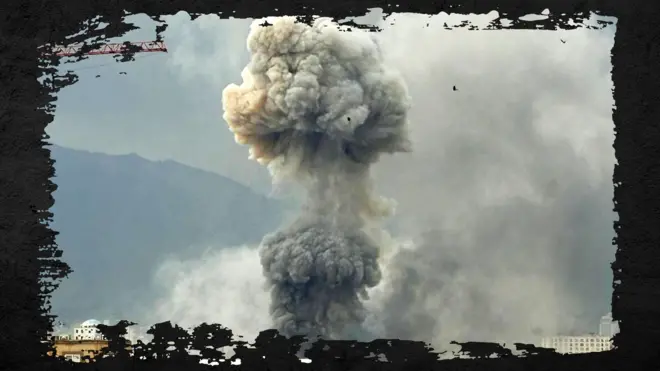 Smoke from an explosion rising in two clouds, one above the other, with two buildings at the bottom of the image and a hillside in the background. The image has had a frame added in the style of distressed black paint.
