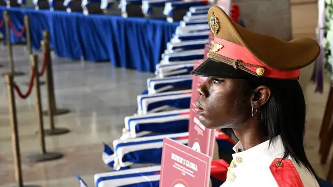 Una mujer uniformada monta guardia al lado de una fila de pequeños féretros con las cenizas de los muertos, cubiertos con banderas cubanas