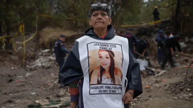 Una mujer luce un peto blanco con una foto de su hija y la leyenda "¿En dónde está Pamela?". En el fondo, hombres trabajan excavando la tierra en un área delimitada por una cinta amarilla. 