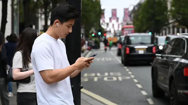 Молодой человек в белой футболке сморит в телефон на переходе через дорогу, на дороге видны машины, красный даблдекер и висящие поперек британские флаги