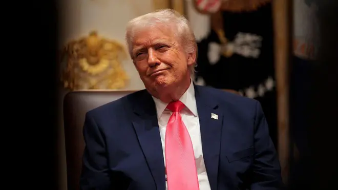 تصویری از دونالد ترامپ، رئیس جمهور امریکا که پشت میز خود نشسته و لبخنی معنی‌دار به لب دارد.