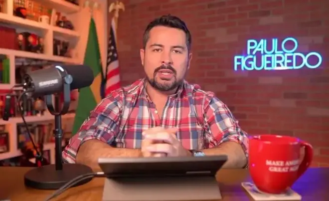 Paulo Figueiredo está em uma mesa com um notebook, um microfone e uma xícara. Atrás há uma bandeira do Brasil e dos EUA e uma logo iluminada com o nome dele. 