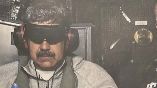 Мадуро уйги кийимида кўзлари қора тўсиқ билан қопланган, қулоқлари ҳам қулоқчин билан беркитилган ҳолда кўринишидан ҳарбий вертолёт ўриндиғида ўтирибди