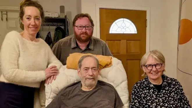 В кресле сидит пожилой седой мужчина с бородой, справа от него пожилая женщина в очках, слева женщина помоложе и за креслом бородатый мужчина в очках. Женщины улыбаются, пожилой мужчина, возможно, тоже, мужчина помоложе серьёзен.