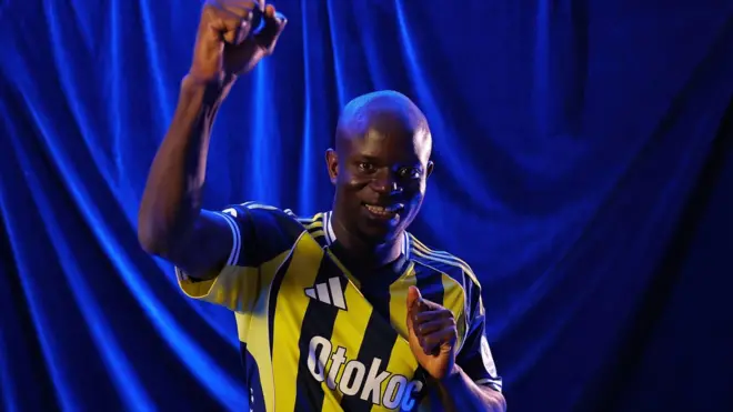Kante Fenerbahçe formasıyla poz veriyor. Havadaki elini yumruk yapmış, yüzü gülüyor.