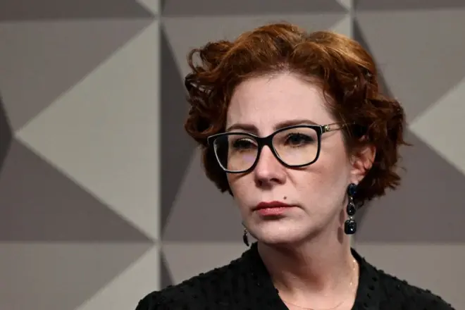 Carla Zambelli olhando para o lado. Ela usa óculos de grau, brincos e uma blusa preta.