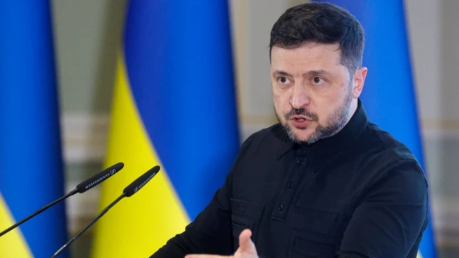 Volodimir Zelenskiy, kürsüde konuşma yapıyor, üzerinde siyah bir kıyafet var. Arkada da bayraklar duruyor.